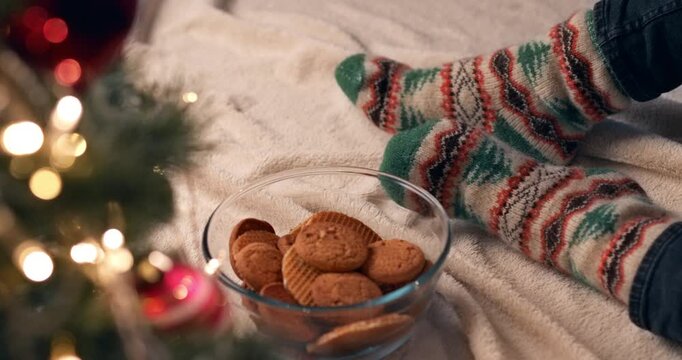 A glass bowl of cookies rests on a soft blanket beside feet in cozy patterned knitted socks with glowing Christmas lights nearby.
