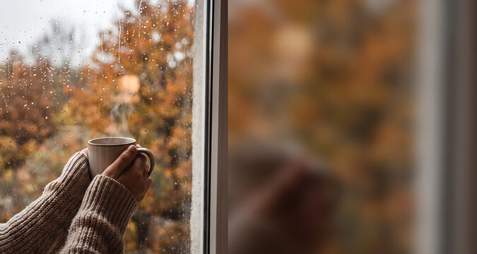 Hands in a cozy sweater holding a warm mug beside a rain-streaked window in autumn