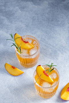 Two glasses hold refreshing drinks with ice and peach slices with a sprig of rosemary on a gray background. Herbs are added for flavor. The scene is bright and set on a light surface.