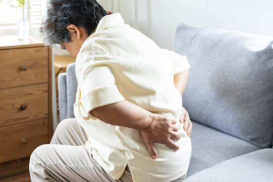 Pain body muscles stiff problem, suffer asian aged woman painful back, symptom injury ache from long sitting, standing, hand holding massaging or rubbing lower back hurt, sore sitting on sofa at home.