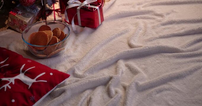 A hand reaches for a bowl of cookies as feet in red Christmas socks rest near a tree with wrapped gifts and a reindeer pillow.