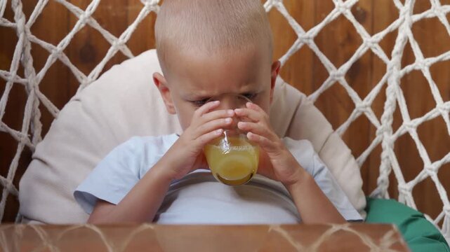 A focused little boy eagerly gulps fresh yellow juice from a clear glass. He sits comfortably inside a cozy macrame net chair against a warm wooden background.
