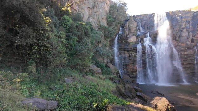 Panning shot revealing Salto Corumba waterfall and tourists from behind dense forest vegetation during dry season