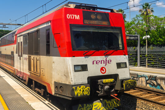 Barcelona, ​​Spain, June 29, 2026. A red and white Renfe double-decker passenger train with yellow graffiti tags on the front bumper sits on tracks beside a station platform.