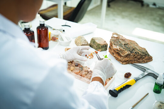 Laboratory scientist rock mineral sample glove, geology research with hammer, bottle, tray, and specimen on workbench during careful analysis in bright science workspace