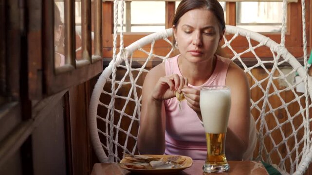 A relaxed woman contentedly cleans dried fish while sitting behind a tall glass of foamy beer. She enjoys her snack peacefully on a wooden balcony next to a white hanging chair.