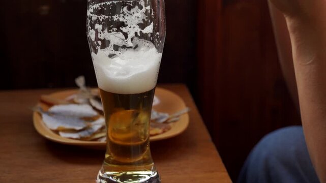 A tall glass of foamy beer stands on a wooden table next to a plate of dried fish. A relaxed person enjoys a peaceful, cozy evening break at home, feeling completely content and satisfied.