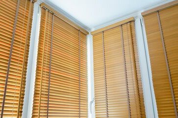 Wooden venetian blinds cover corner windows with white PVC frames in a bright sunlit room interior © Arseniy