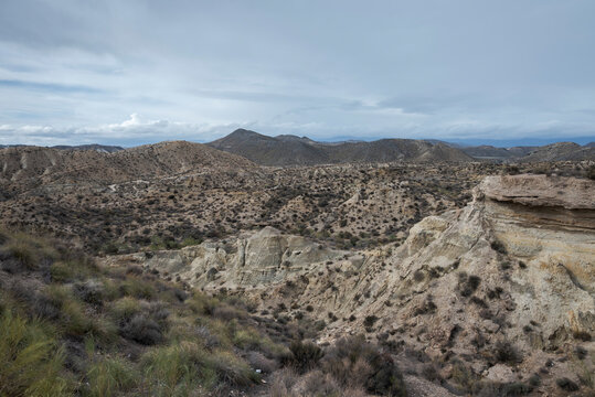 The arid landscape of Tabernas Desert in Almeria, Spain, with rugged hills, sparse vegetation, and rocky outcrops under a cloudy sky. It&rsquo;s a popular filming location