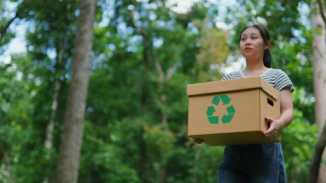 Young Asian woman volunteer holding a recycling box in the park. Concept of recycling, environmental protection, sustainability, zero waste, and ESG strategy on green natural forest background.