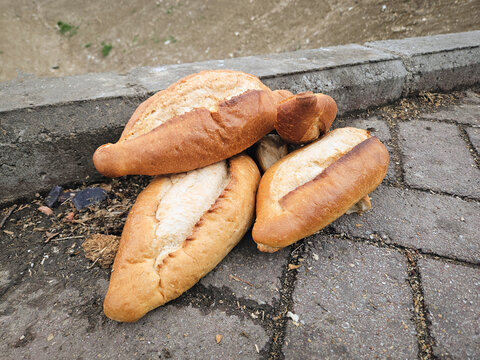 Pile of fresh bread loaves discarded on a paved city sidewalk next to a curb, with a blurred urban street background