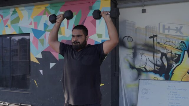 Bearded South Asian male athlete performing a standing dumbbell shoulder press during a strength training workout in a modern indoor fitness studio. Camera tracks in and then tracks out during the exe