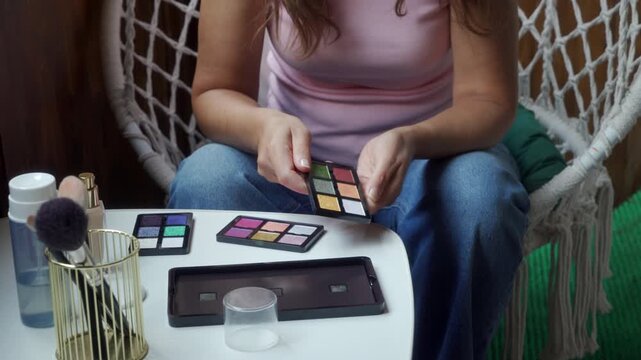 An inspired woman enthusiastically chooses colors from a small magnetic eyeshadow palette block. She sits comfortably at a white table on a cozy balcony next to a white macrame chair.