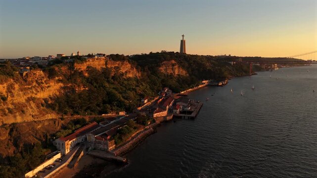 The drone glides forward along the Almada coastal waterfront with the Cristo Rei statue rising above the cliffs ahead.