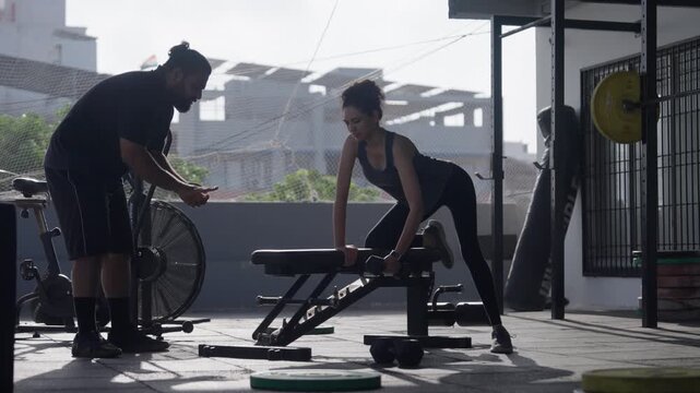 Wide shot of a male gym coach assisting and instructing a young woman doing dumbbell single arm row exercise on a bench.