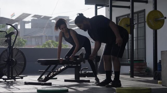 Wide shot of a bearded muscular man performing single arm dumbbell rows leaning on an exercise bench in a gym setup.