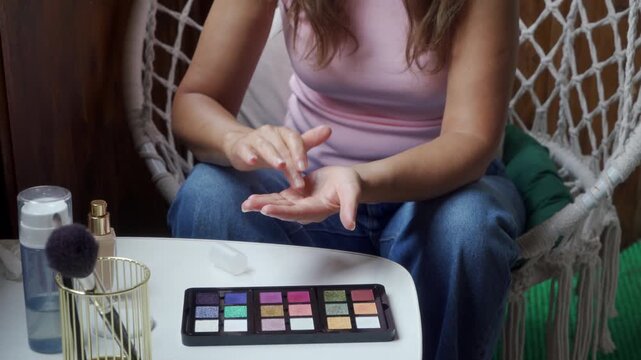 A relaxed woman happily blends a makeup product into her hand with quick movements. She sits contentedly by a small white table with cosmetics on a cozy balcony next to a white macrame chair.