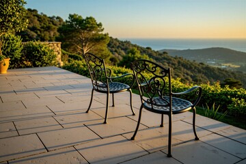 Obraz premium Two metal chairs sit on stone terrace at sunset. Furniture faces scenic ocean view over green hills. Outdoor space provides calm atmosphere for relaxation on luxury resort patio. Empty seats wait for