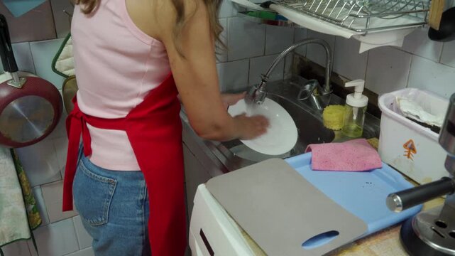 A determined woman contentedly scrubs a white ceramic plate with a sponge. She works smoothly at a metal kitchen sink surrounded by tools on a busy, tiled counter.