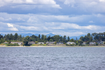 Fototapeta premium Waterfront Homes Along Sidney Shoreline With Mountains And Cloudy Sky on Vancouver Island BC