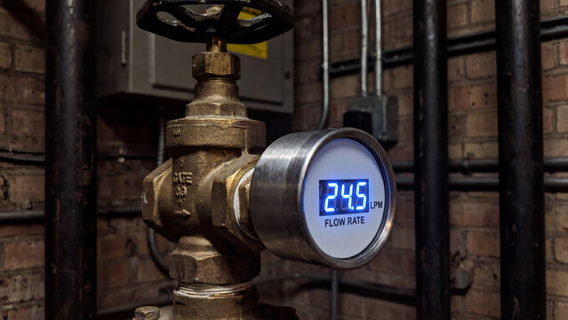 Industrial flow meter displays a digital reading against a backdrop of weathered pipes and brick walls.
