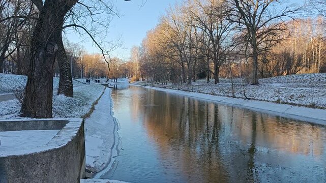 A channel partially covered with ice runs between the willows and birches, with concrete banks dusted with snow. Cars are driving, people are walking along the path. Sunny winter weather and blue sky