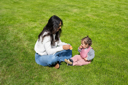 Mother Feeding Baby Girl While Sitting On Green Lawn In London
