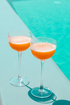 Two orange cocktails sit by a turquoise pool in bright sunlight, evoking a relaxed summer vacation vibe.