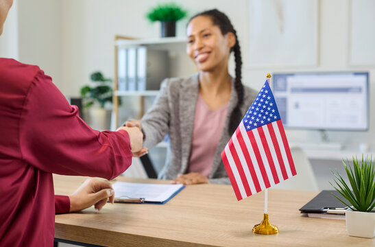 Immigration consultant client handshake approval visa interview documents. Advisor welcomes applicant in office with USA flag, indicating successful application. Trust and approval theme shown.