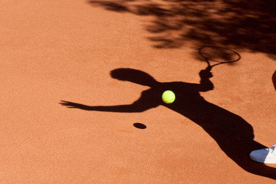 Shadow Tennis Player Hits Ball on Clay Court During Warm Afternoon