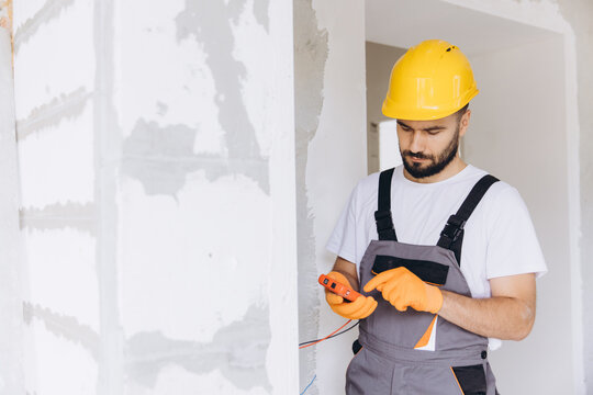 Young man in a hard hat and protective gloves, performing electrical installation and repair on a construction site