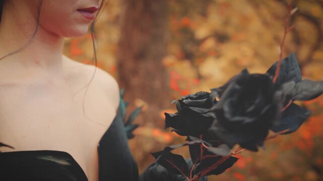 close up shot part of face mouth lips, gothic woman dark queen holding black rose flower in hands long gloves stroking petals. Happy fantasy sexy girl in autumn nature red leaves forest trees park 4k.
