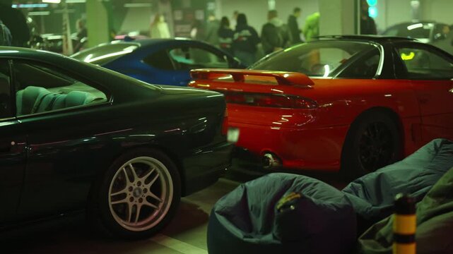 Modern sports cars parked in an underground parking garage during a night-time car exhibition with people walking in the background, showing off tuning, spoilers, and custom wheels