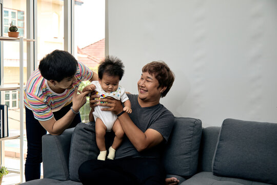 Two smiling men holding and supporting a baby beside a sofa at home, sharing care, affection, bonding, happiness, tenderness, comfort, playful interaction, and loving togetherness