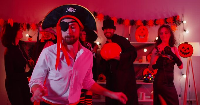 Joyful young man in pirate costume celebrating at Halloween party with friends. Group of people in spooky outfits dancing, having fun in decorated room with festive lights and pumpkin decorations.