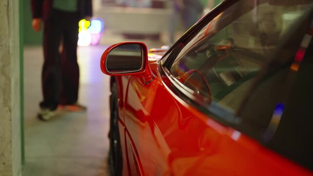 Close up view of a luxury red sports car parked in an underground garage with people walking past in the blurred background and colorful lights reflecting on the shiny surface of the car