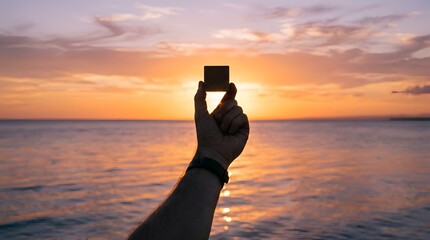 Fototapeta premium Silhouetted hand holding dark cube raised triumphantly against glowing golden sunset over serene ocean waters, tranquil beach evening with warm sky and reflective horizon