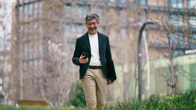 Video of happy mature businessman using his phone while walking through the park