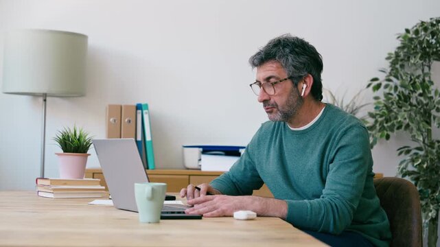 Video of mature man working with laptop while taking notes at the office