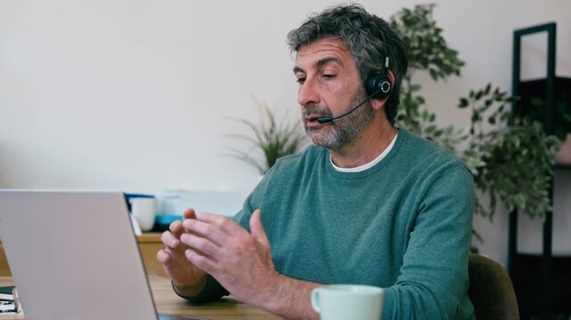 Video of mature man doing a videocall with laptop while talking with earbud at the office
