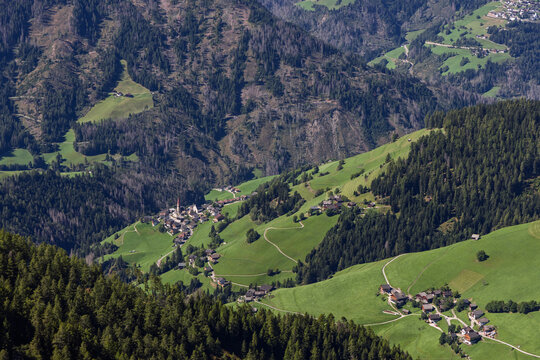 Pieve di Marebbe and scattered farmsteads sit across vivid green slopes and dark forest, viewed from high on Kronplatz in South Tyrol.