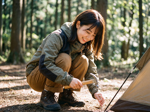 森の木漏れ日の中で笑顔でテントのペグを打つソロキャンプの女性