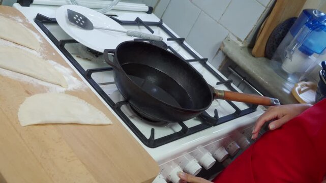 A woman turns the knob on a gas stove to heat oil in a pan. She feels focused, excited, and eager to fry the prepared pastries waiting on the nearby wooden board.