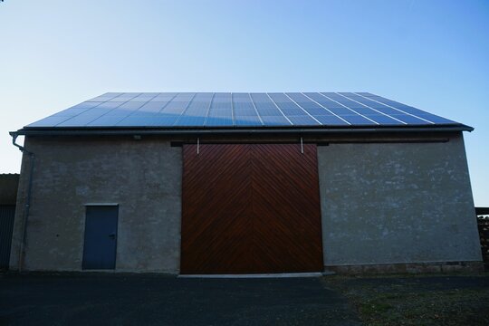 Gro&szlig;e alte sanierte Bauernscheune mit blauer T&uuml;r, rotem Holztor und blauen Solarpanels vor blauem Himmel bei Sonne am Abend im Sommer