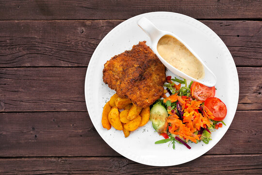 Classic German Jagerschnitzel with a mushroom cream sauce, potato wedges and salad. Overhead view on a dark wood table background.