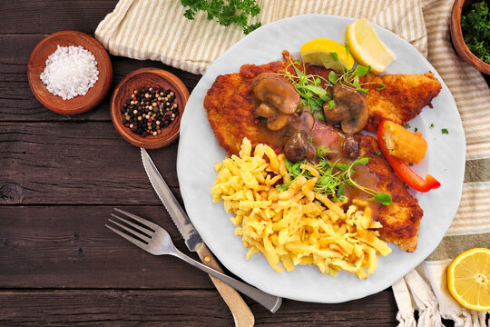 Classic German Jagerschnitzel with a brown mushroom gravy and spaetzle noodles. Top down view table scene against a dark wood background.