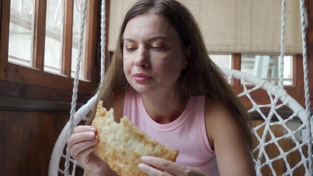 A thoughtful woman holds a bitten golden pastry while sitting in a hanging swing chair on a cozy porch. She looks relaxed and content as she enjoys her delicious homemade snack.