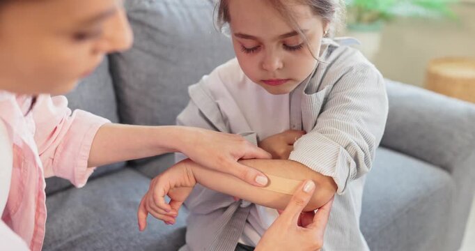 Child injury arm elbow bandage plaster caregiver care. An adult applies an adhesive strip to a sore elbow on a sofa at home, offering reassurance. Concept is caregiving and child safety.