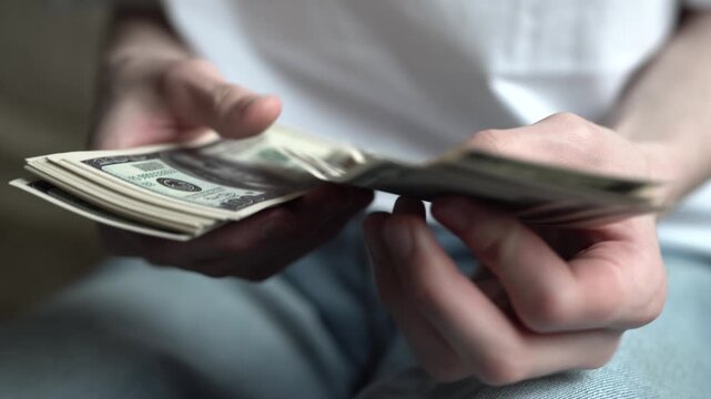 A man is counting a stack of money with hands. The man carefully handles the cash, examining each bill closely in a low-lit setting.