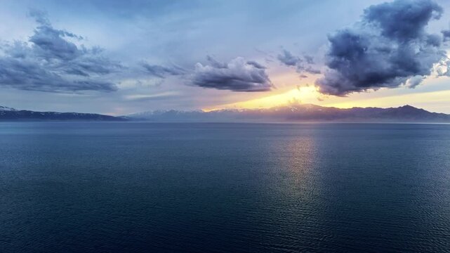 Aerial view of dramatic golden sunset over a vast blue lake framed by distant snow-capped mountains and moody storm clouds.